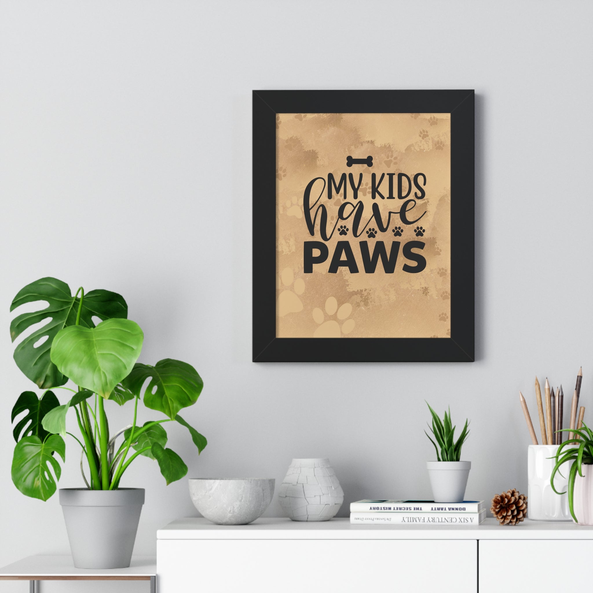 Framed print with 'My kids have paws' text on a wall above a white cabinet with plants and decor items.