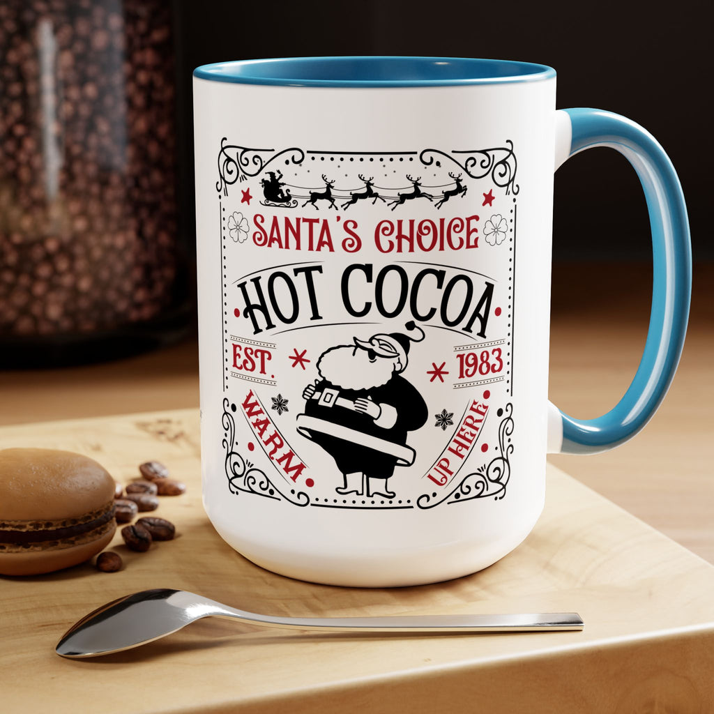 White mug with blue interior and handle, featuring 'Santa's Choice Hot Cocoa' design, on a wooden surface with coffee beans and a spoon.