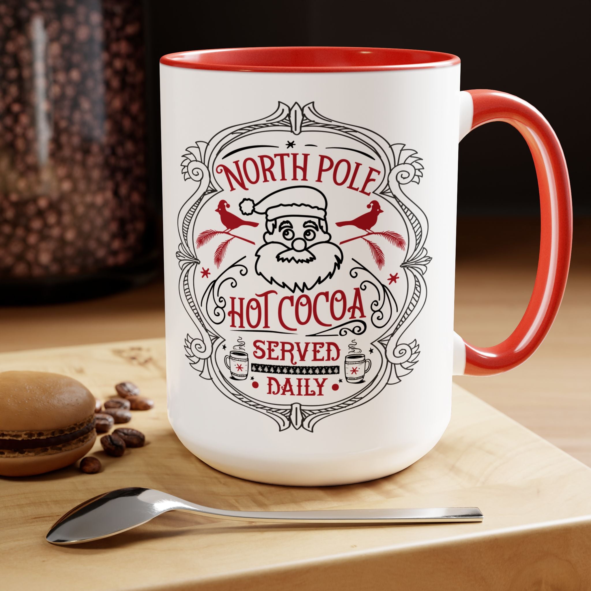 White mug with red interior and handle, featuring a Santa Claus design and text, on a wooden surface with coffee beans and a spoon.
