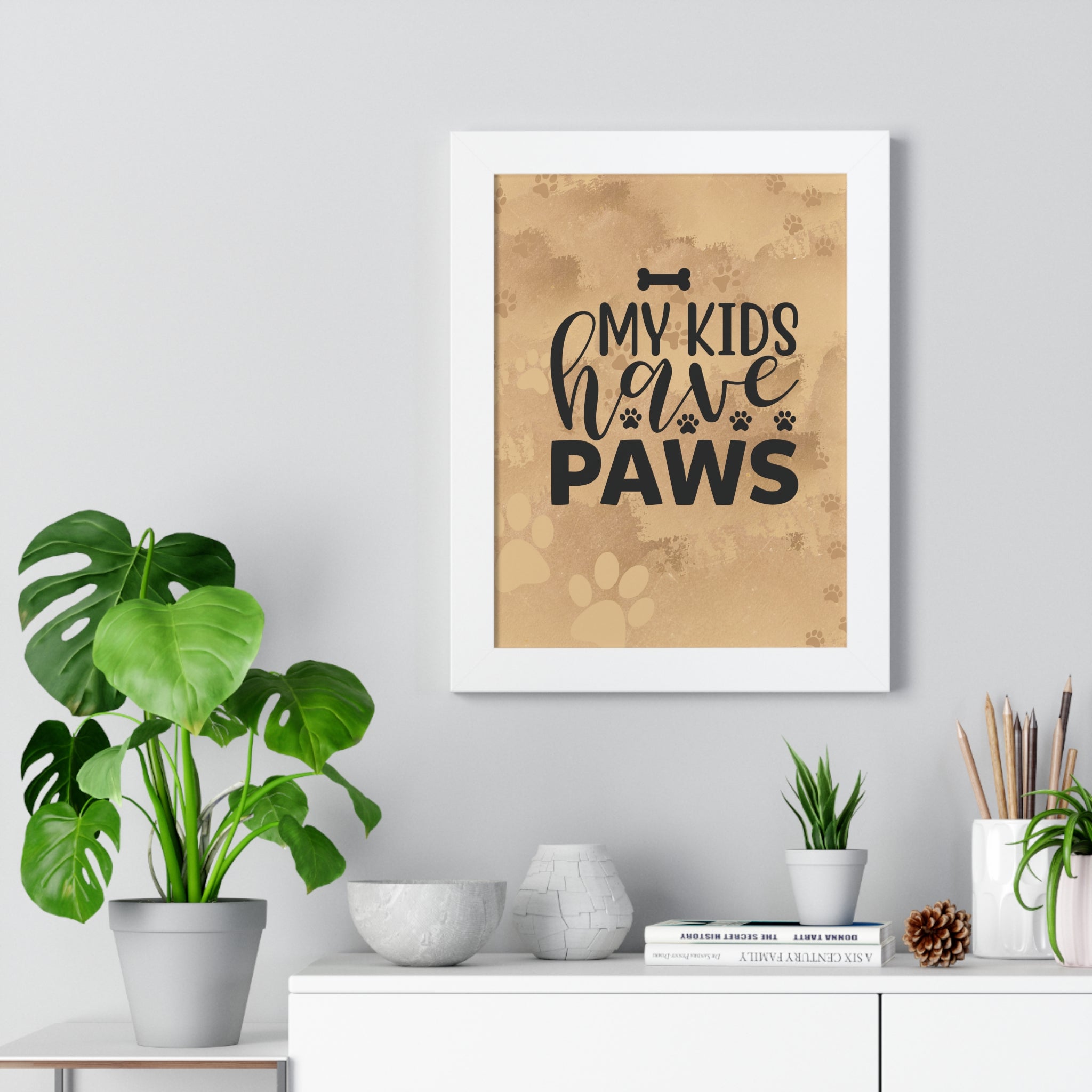 Framed poster with 'My kids have paws' text on a wall above a white cabinet with plants.