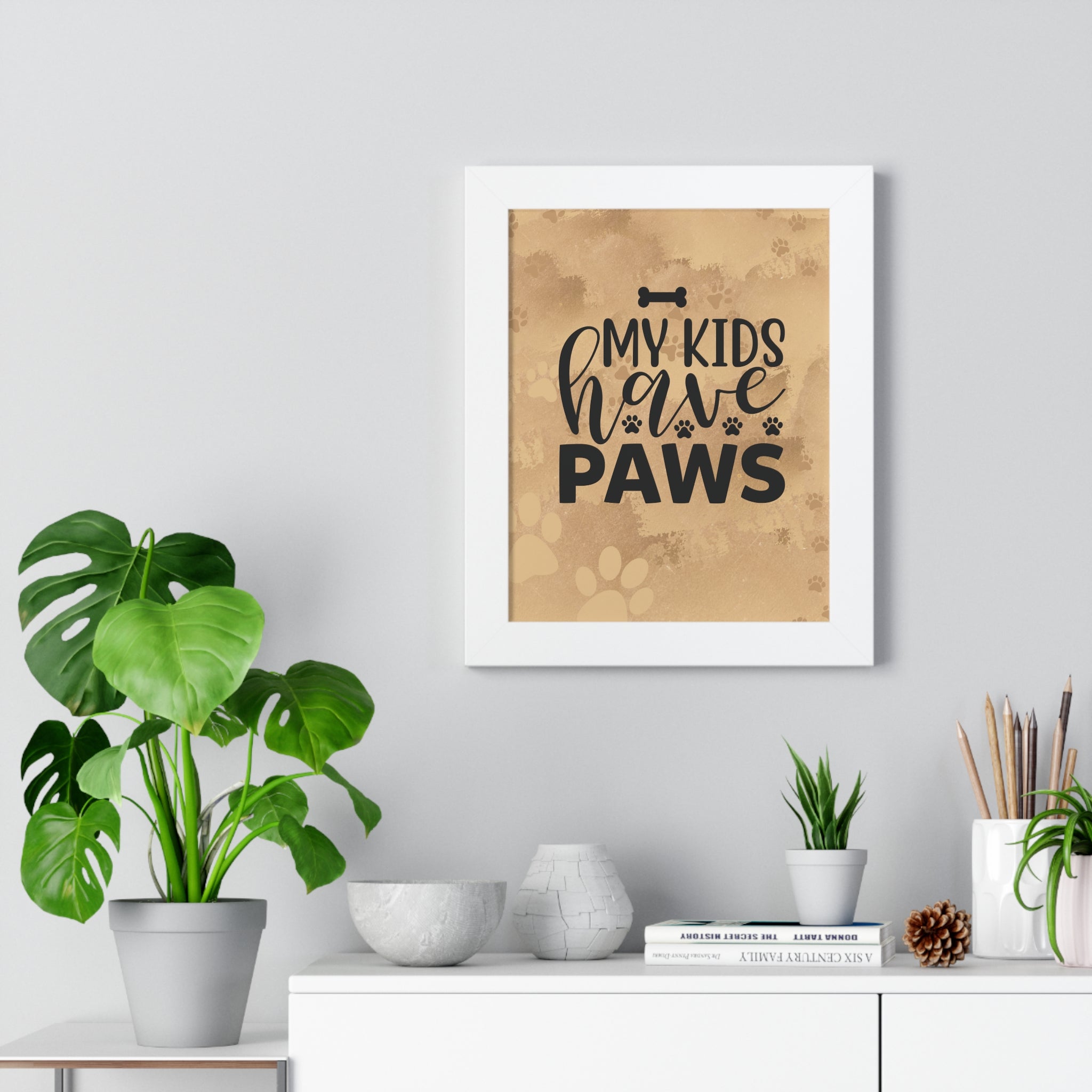 Framed print with 'My kids have paws' text on a wall above a white cabinet with plants.