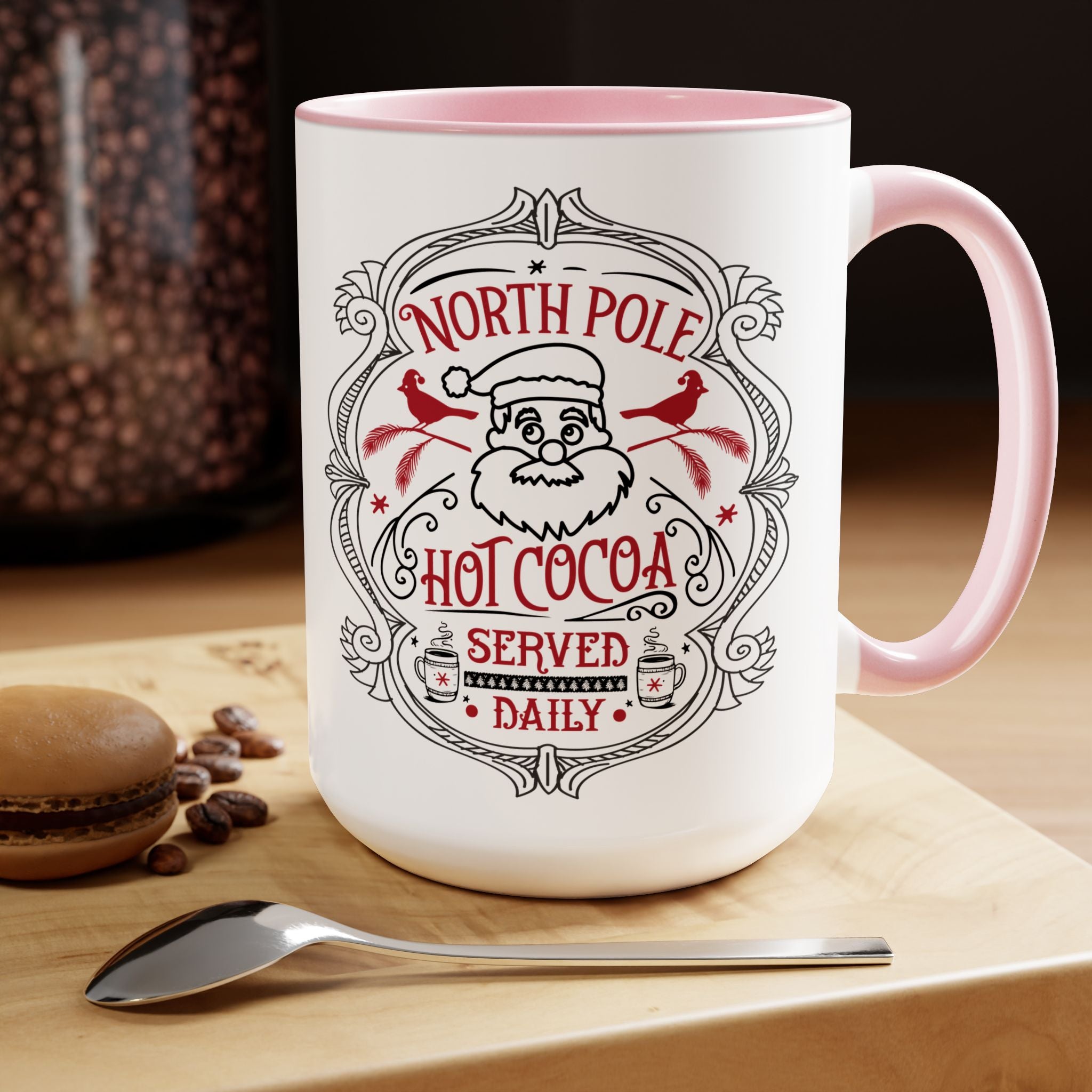 White mug with pink interior and handle, featuring a Santa Claus design and text, on a wooden surface with coffee beans and a spoon.
